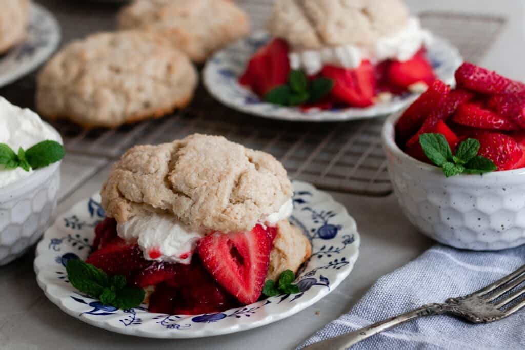 two sourdough shortcakes with strawberries and whipped cream on blue and white plates with a bowl of strawberry in a white bowl to the right
