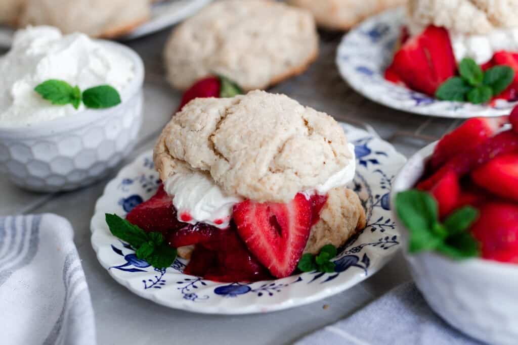 sourdough shortcake with sweetened strawberry sauce and homemade whipped cream sandwiched between the shortcakes. Whipped cream in a white container sits right behind the shortcakes and a bowl of strawberries to the right