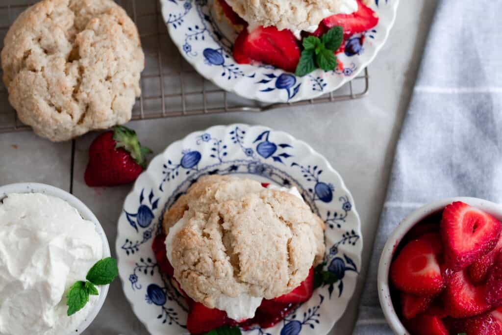 overhead photo of strawberry shortcakes on a blue and white floral plates with whipped cream and strawberries. A bowl of strawberries to the right of the biscuits and a sourdough biscuit and bowl of whipped cream to the left
