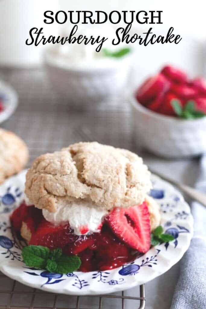 sourdough shortcake biscuit cut in half and layered with strawberries and homemade whipped cream on a blue and white floral plate. One white bowl of sliced strawberries and another white bowl of whipped cream are in the background