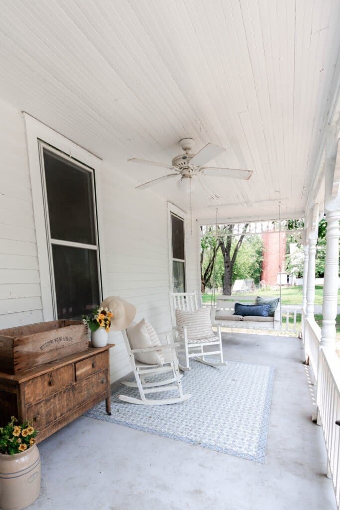 summer farmhouse front porch complete with an antique dresser with a vase of sunflowers, two white rocking chairs, and a porch swing