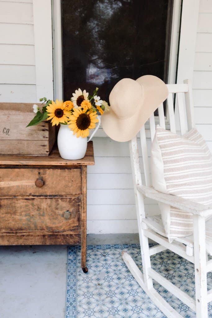 antique dresser on a front porch with a vase of sunflowers. A rocking chair with a stripped pillow and a hat sitting on top of a blue rug is placed to the right of the dresser