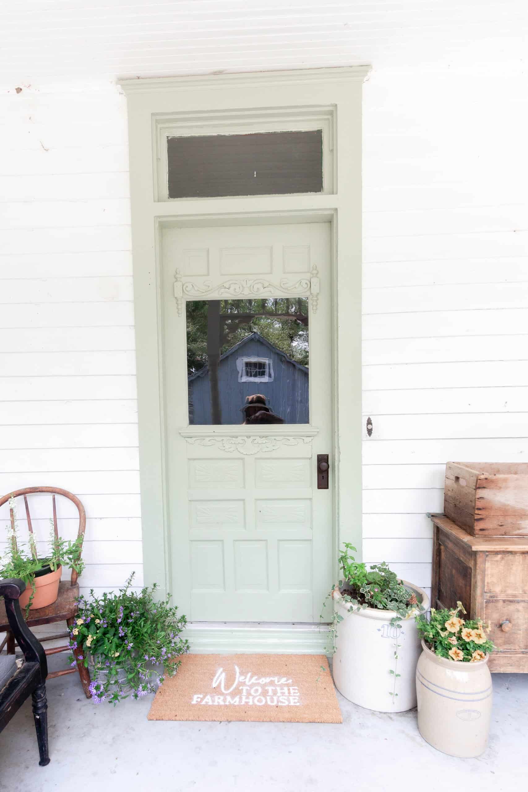 sage green farmhouse front door with a DIY Cricut doormat. Crocks with flowers sit to the right and a antique wooden chair with flowers in a pot on the right of the door