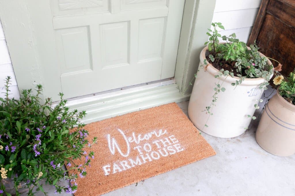 "Welcome to the farmhouse" doormat in front of a sage green antique door with a antique crock full of flower to the right