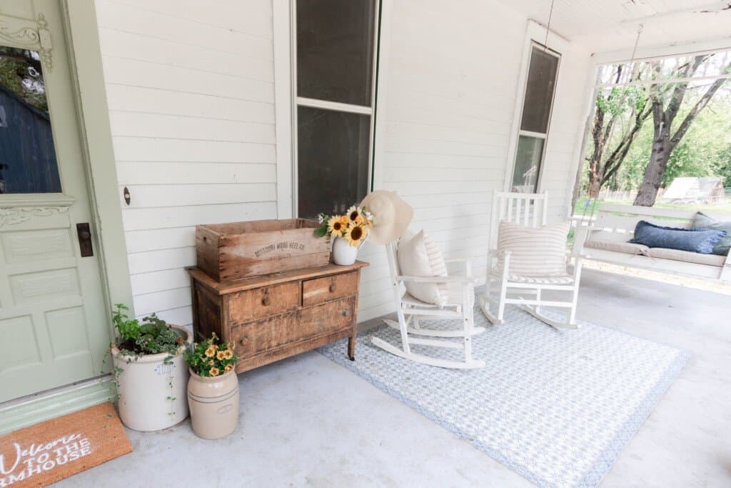 White farmhouse front porch with a sage green door and custom doormat. Antique furniture and white rocking chairs finish off the space