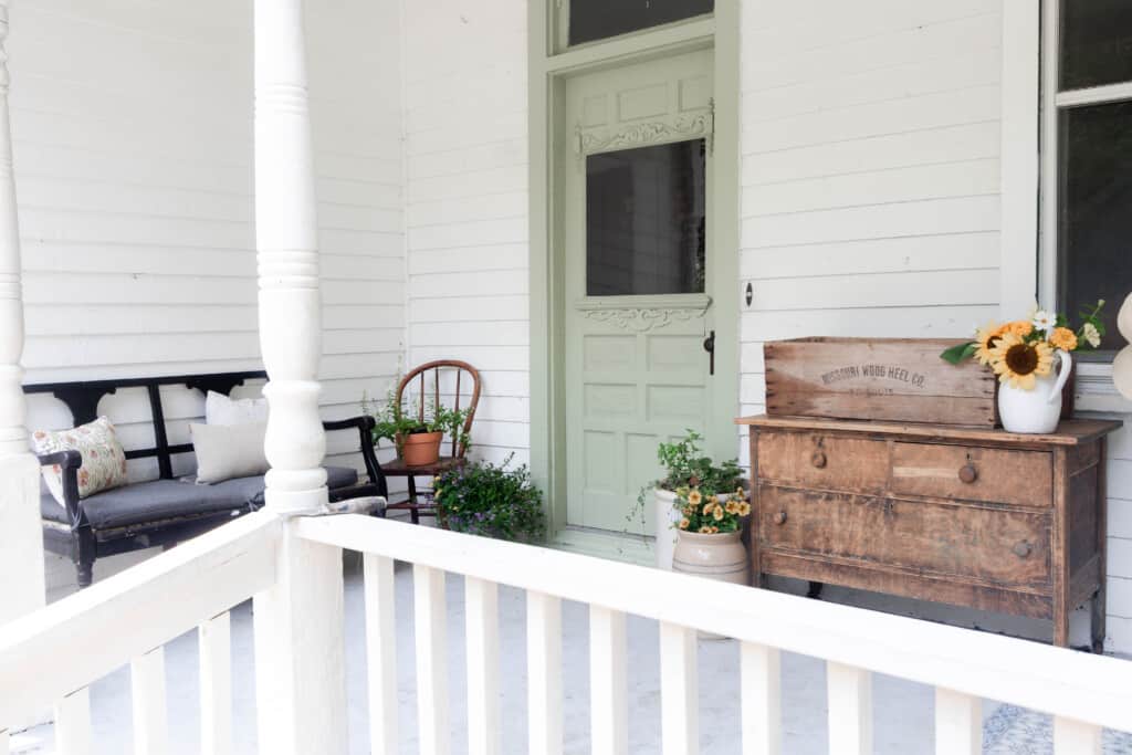 farmhouse front porch with a sage green door, with a antique dresser to the right and a black antique chair to the left