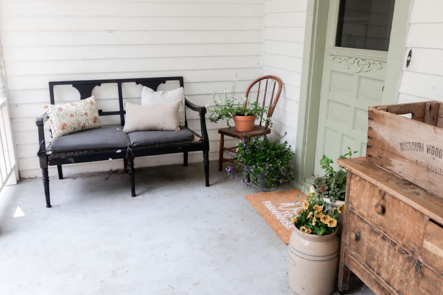 black antique chair with a blue cushions and with pillows. A wooden chair to the right with a pot filled with greenery.