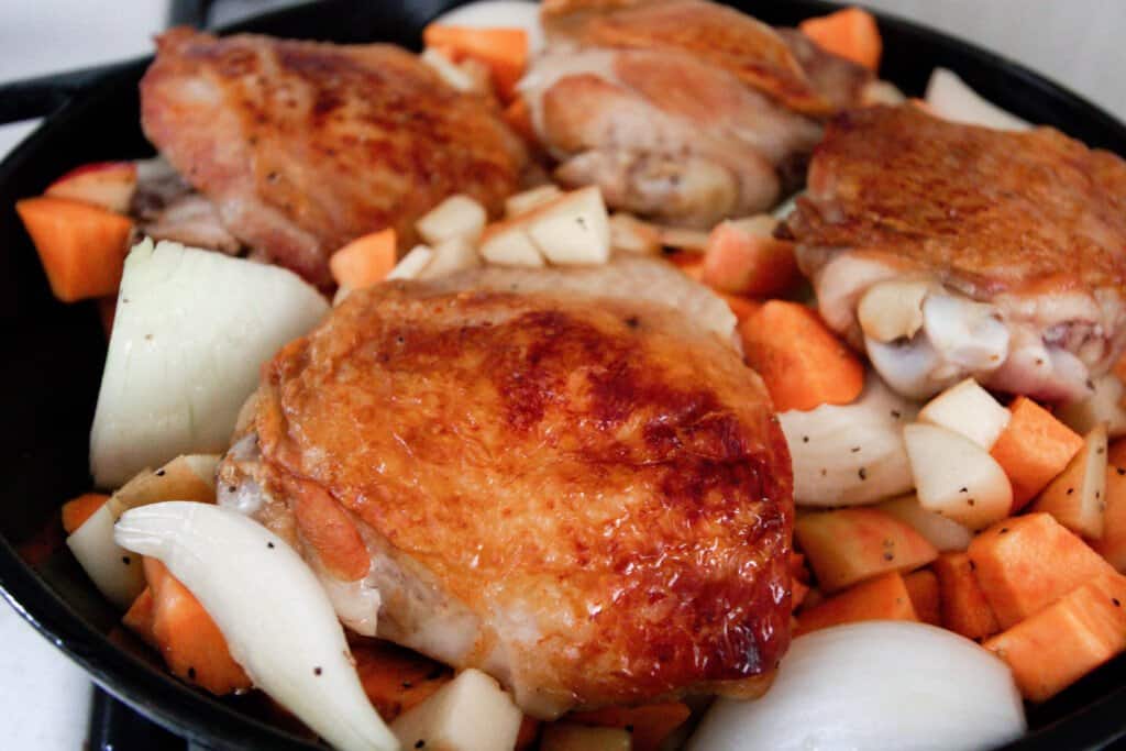 browned chicken thighs in a cast iron skillet with apples, sweet potatoes, and onions arranged around the chicken