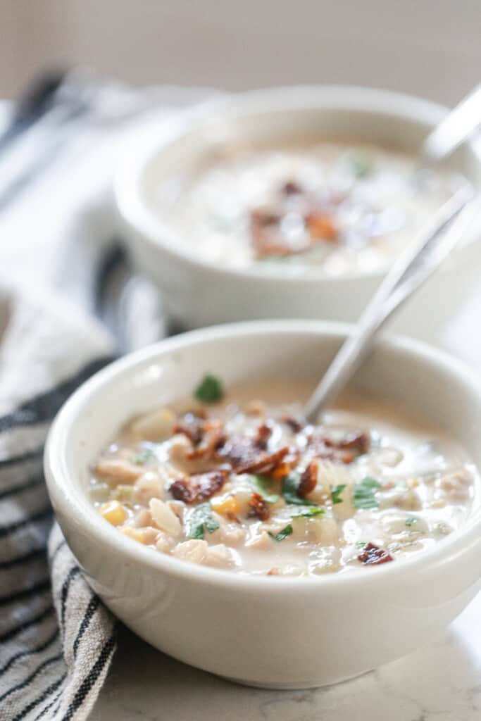 two bowls of chicken and corn chowder topped with fresh herbs and bacon on a white quartz countertop with a black and white stripped towel to the left