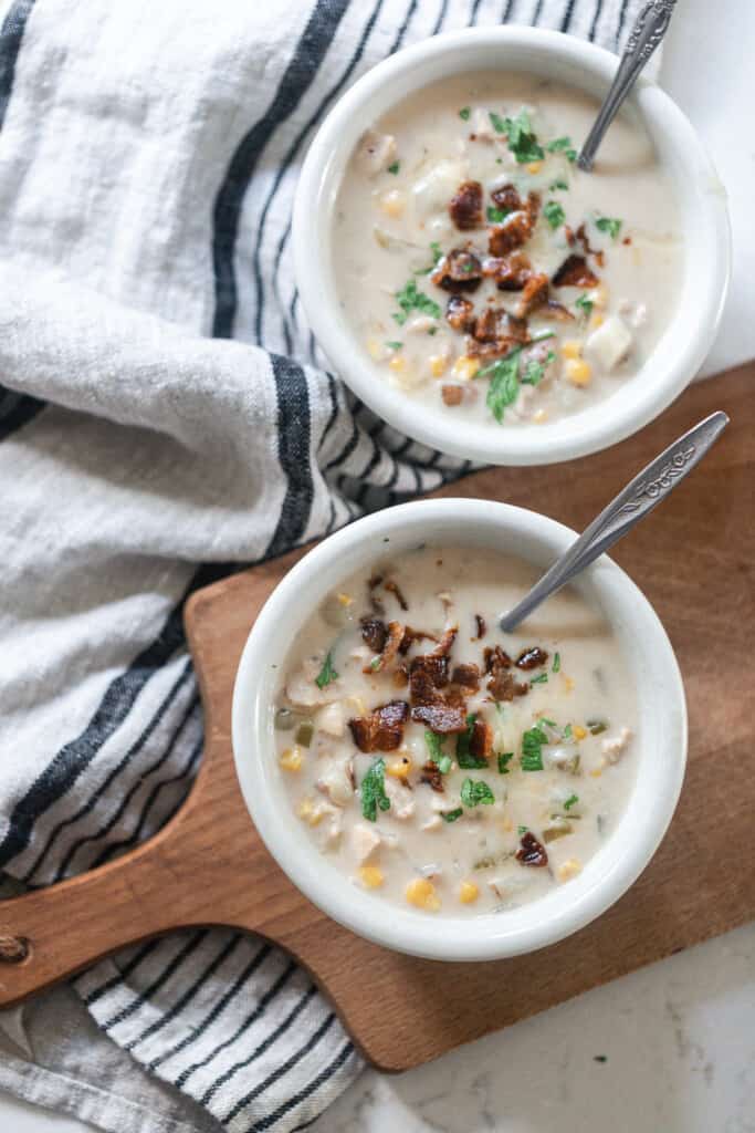 overhead photo of two bowls of creamy corn and chicken chowder. One bowl is on a wood cutting board and another on a white and black towel