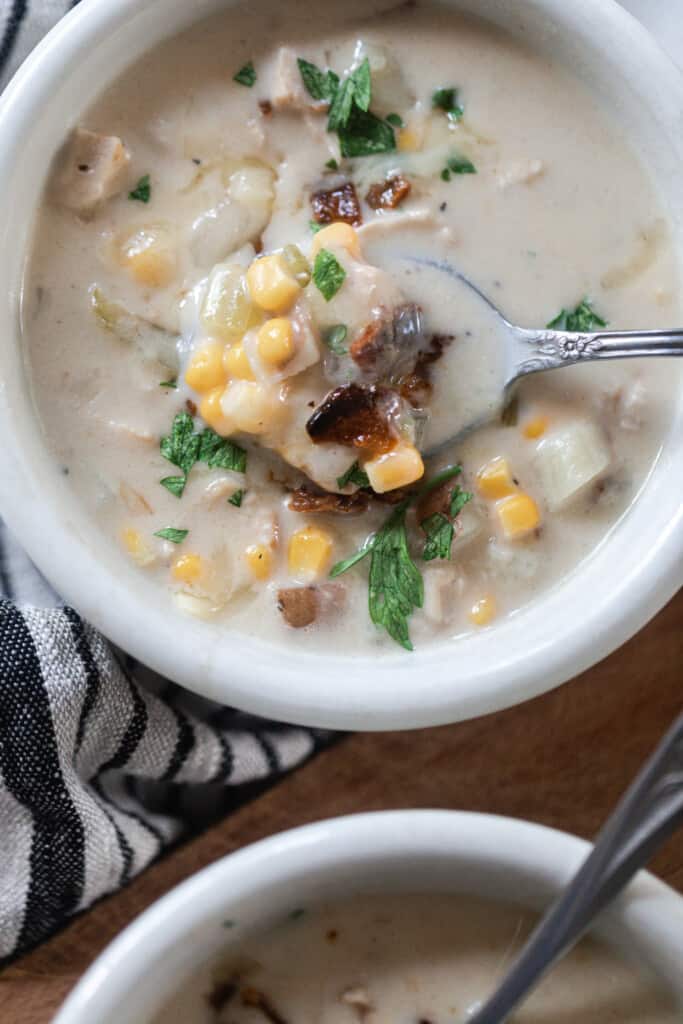 white bowl of creamy chicken and corn chowder with bacon with a spoon in the bowl of soup. Another bowl of soup is right in front resting on a cutting board