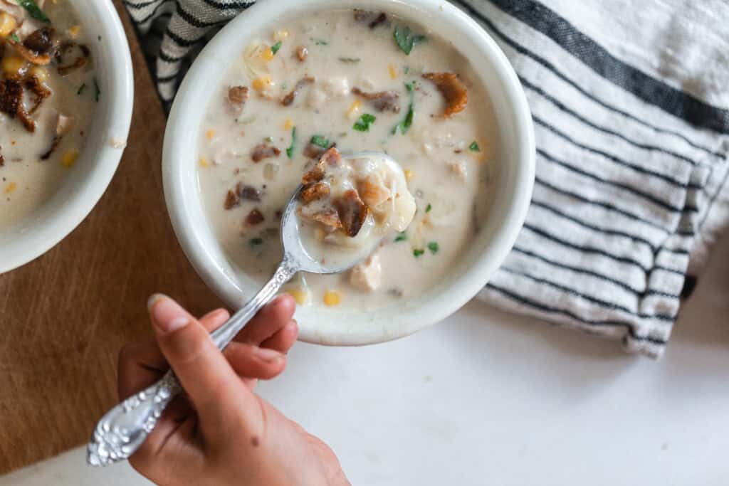 bowl of chicken corn chowder topped with fresh herbs and bacon in a white bowl with a hand holding a spoon taking a spoonful of soup out. The bowl sits on a black and white stripped towel on a wood cutting board