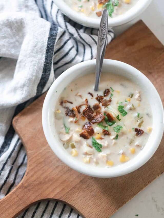 overhead photo of chicken corn chowder topped with bacon and fresh herbs on a wood cutting board on a black and white stripped towel