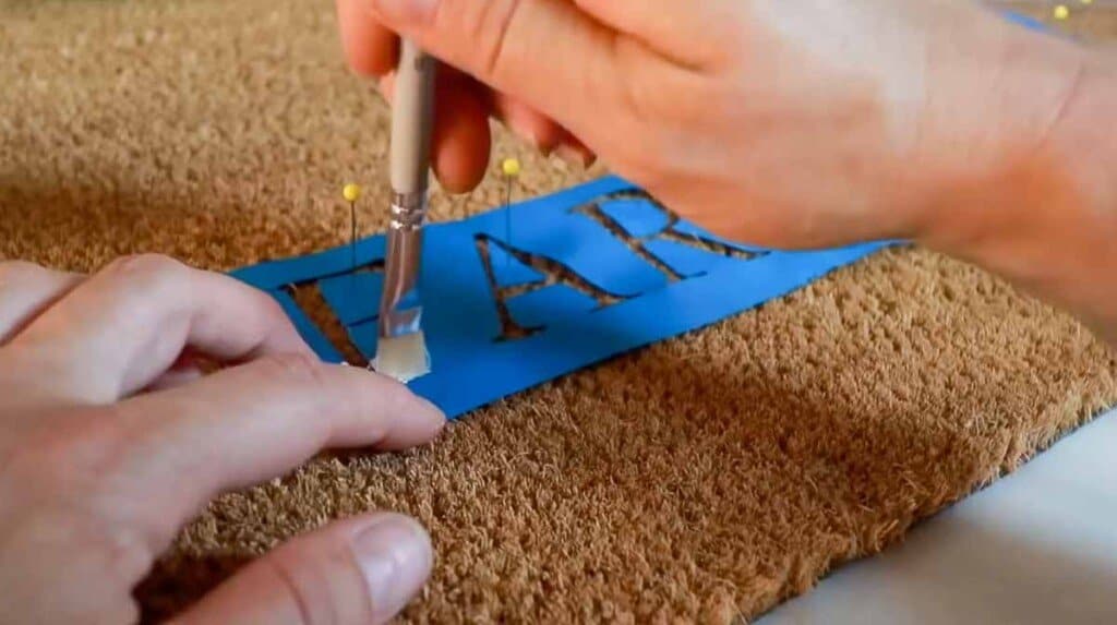 hands using a paintbrush to pain on a blue stencil on a welcome mat