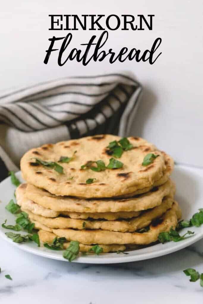 einkorn flat bread stacked with herbs sprinkled over on a white plate with a gray and black stripped tea towel in the background