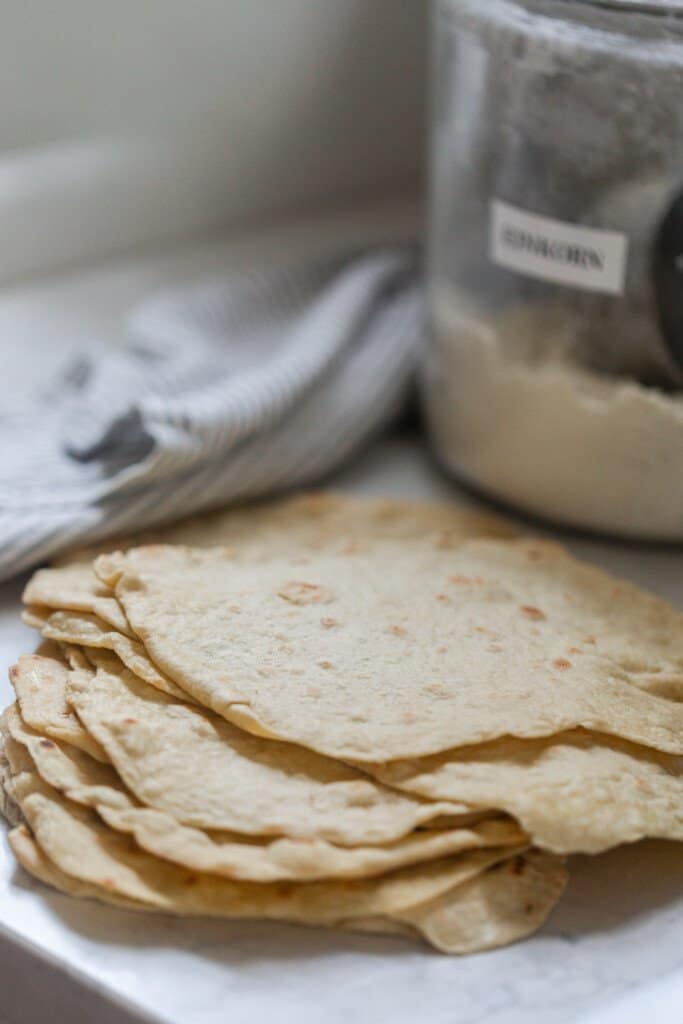 einkorn tortillas stacked on a white quartz countertop with a glass jar of flour and a white and gray towel in the background