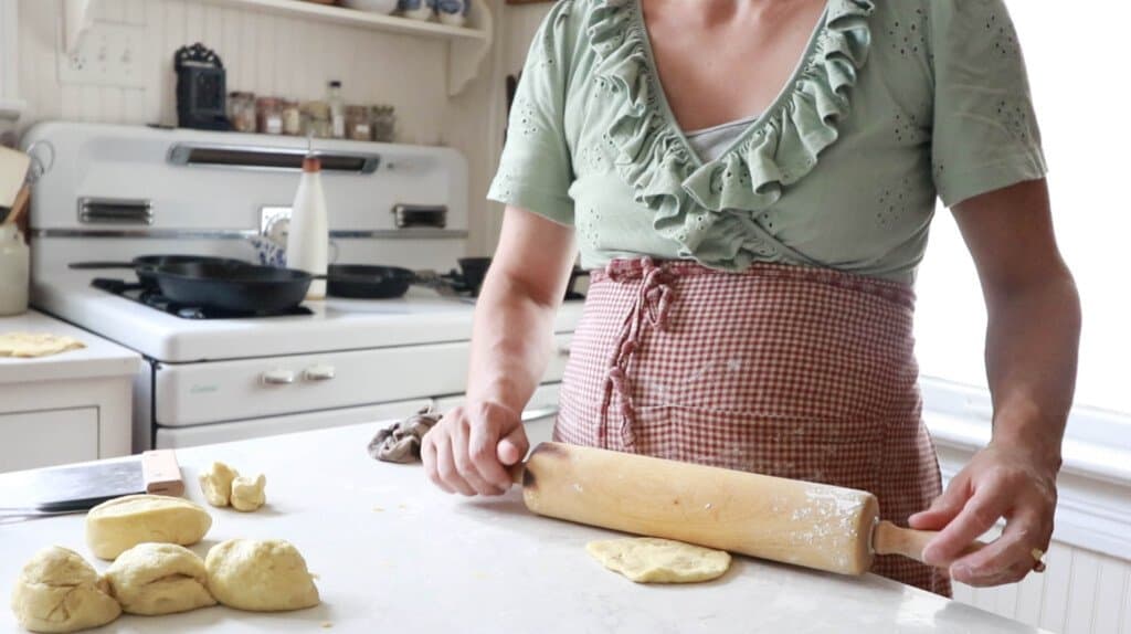 women wearing a light green shirt and red apron rolling out dough with a rolling pin on a white quartz countertop
