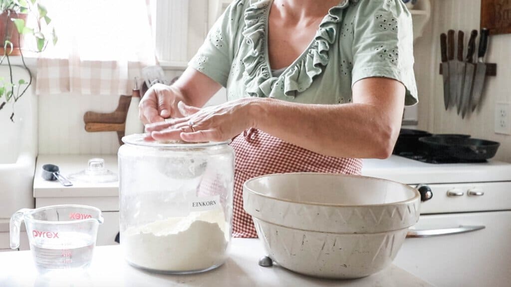 women measuring flour out of a glass jar. A large cream colored bowl sits to the right