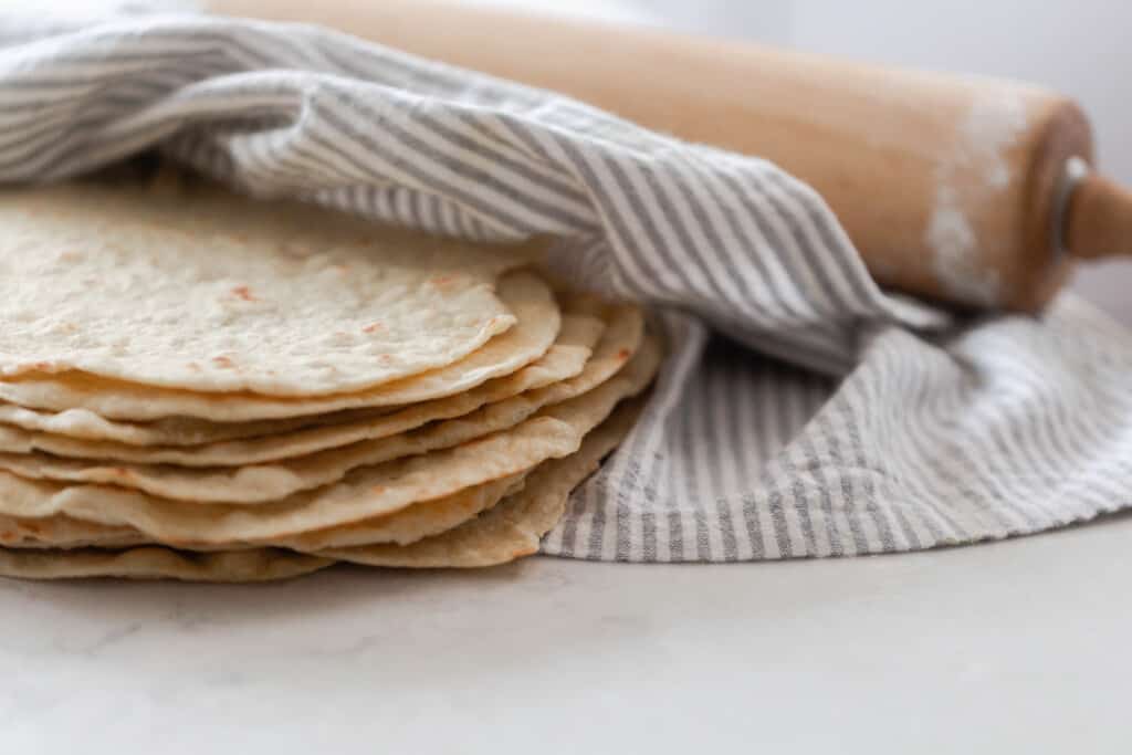 homemade einkorn tortillas sicken up and wrapped with a white and gray towel with a rolling pin in the background