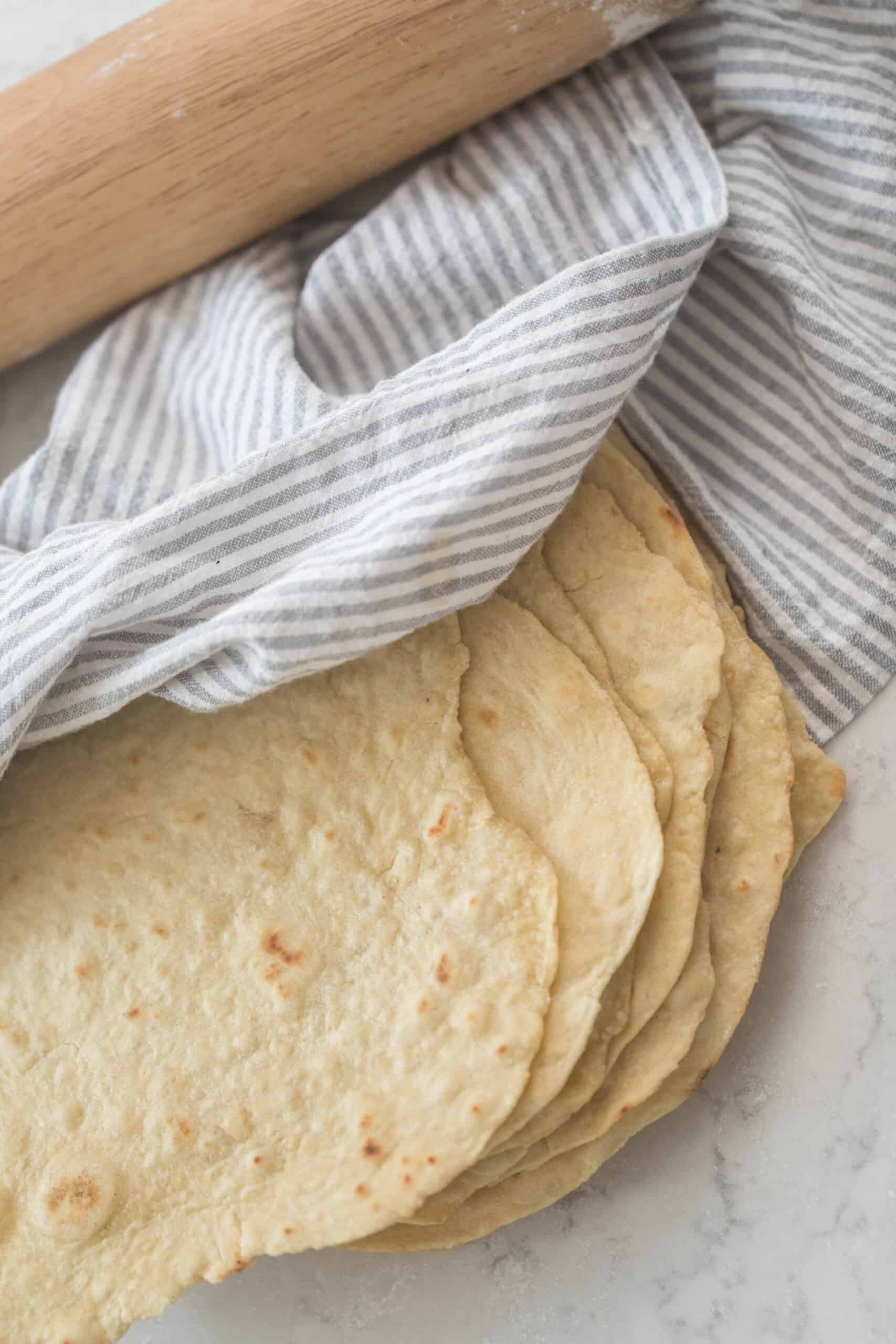 homemade einkorn tortillas stacked on a white quartz countertop. a gray and white towel is draped over the tortillas and a rolling pin is in the background