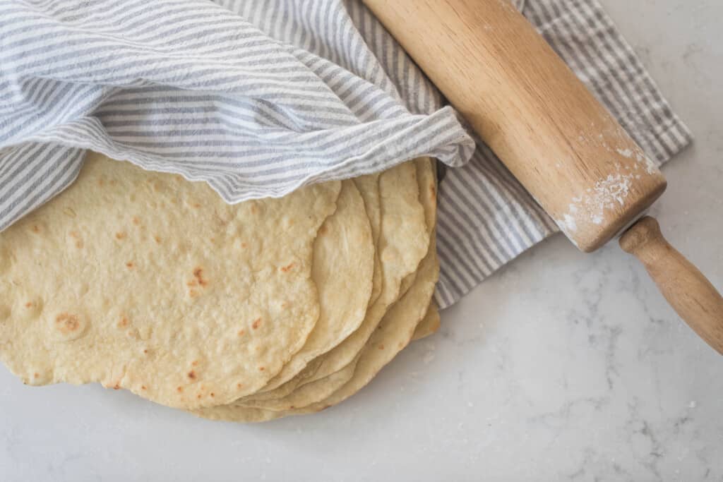 homemade einkorn tortillas stacked on a white quarts countertop and draped with a white and gray stripped towel. A rolling pin rests on top of the towel to the right of the tortillas
