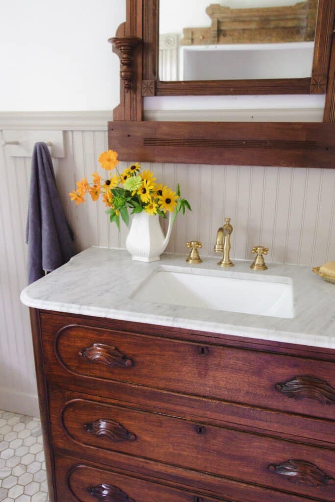 1800's farmhouse bathroom with a antique marble top dresser turned into a bathroom vanity with brass fixtures. A antique mirror hangs above the sink