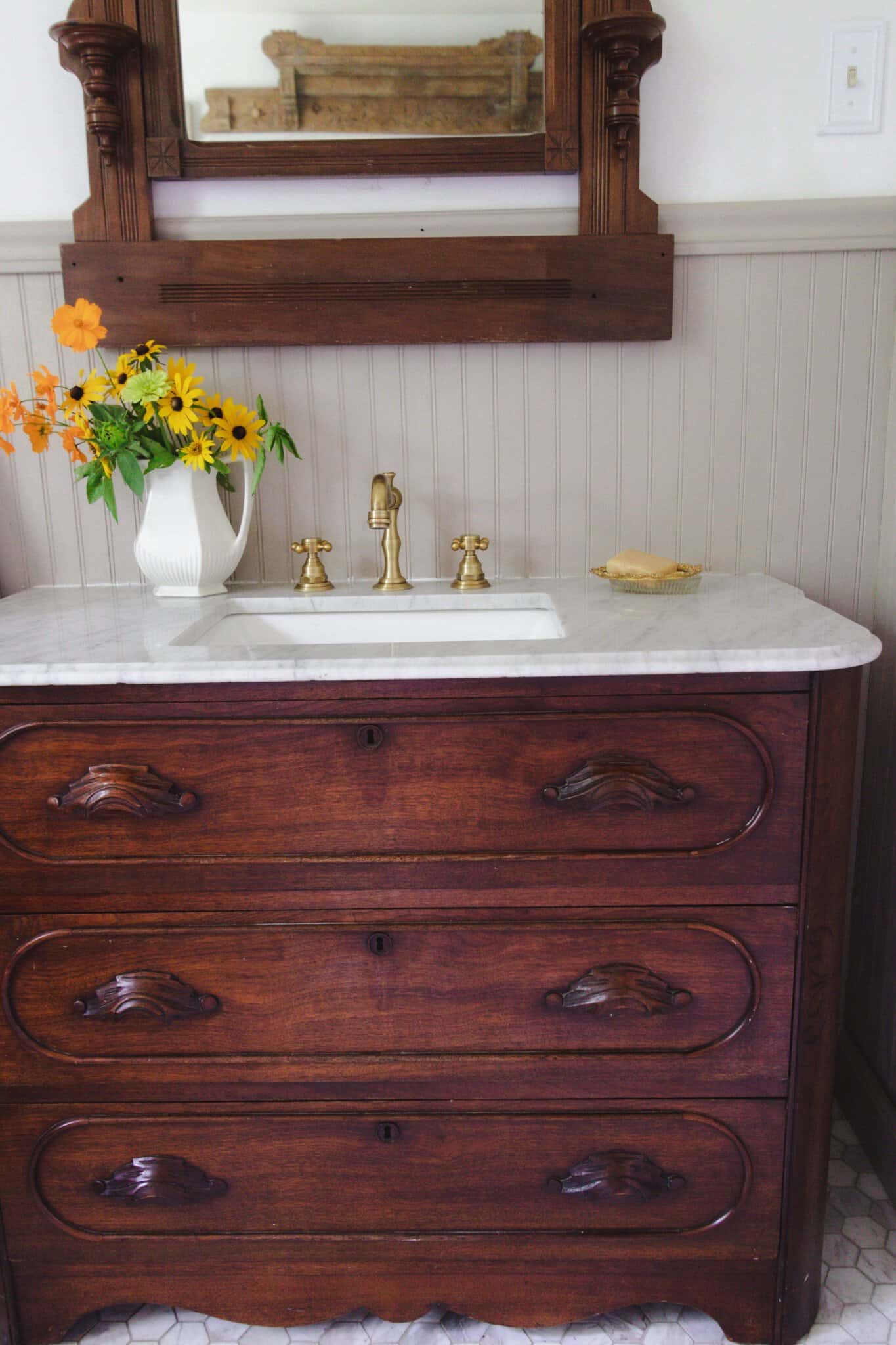 an antique dresser turned into vanity with marble countertop, brass faucets, and flowers.