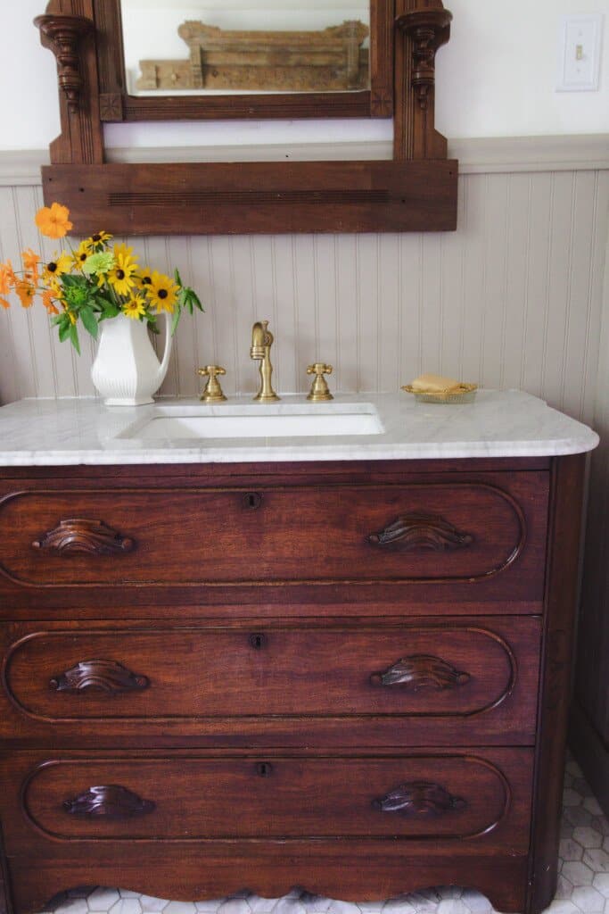 an antique dresser turned into vanity with marble countertop, brass faucets, and flowers.