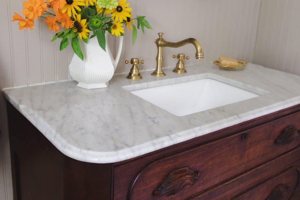 side view of a wood dresser with marble top turned into a vanity with brass faucet. A vase of flower sits to the left of the sink and a brass soap dish to the right