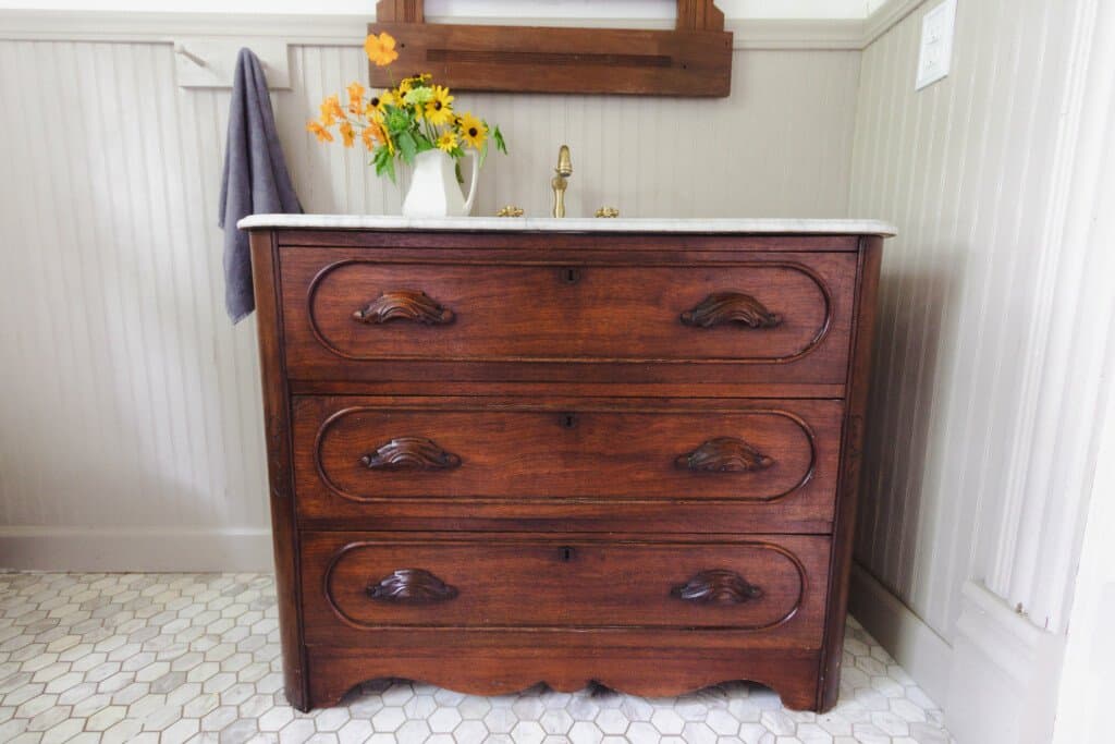 antique dresser turned into a bathroom vanity with a marble top with brass faucet. A vintage cast with flowers sit on the marble counter and a towel hanging on the wall