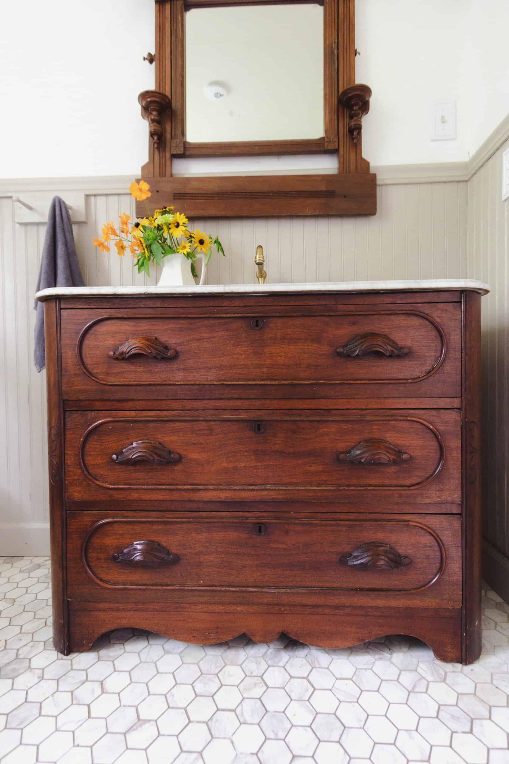 stained wooden antique dresser turned into a bathroom vanity with a marble top. A antique mirror hangs over the vanity.
