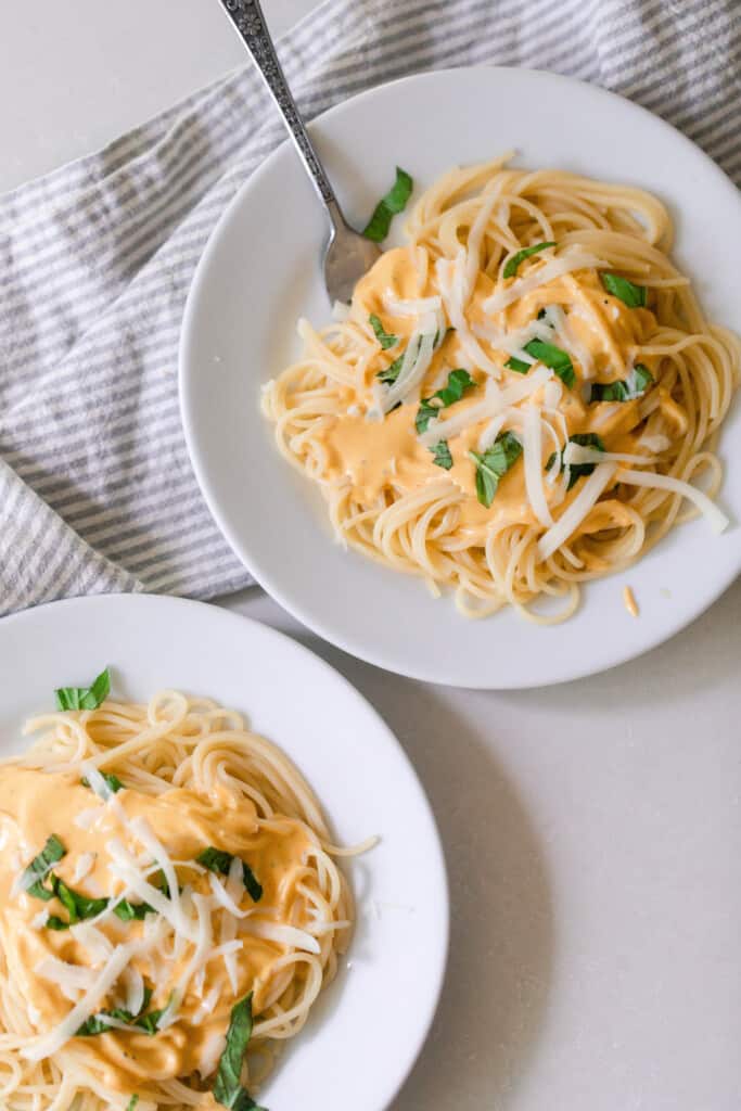 two plates of spaghetti with pumpkin pasta sauce topped with cheese and basil on a white countertop 