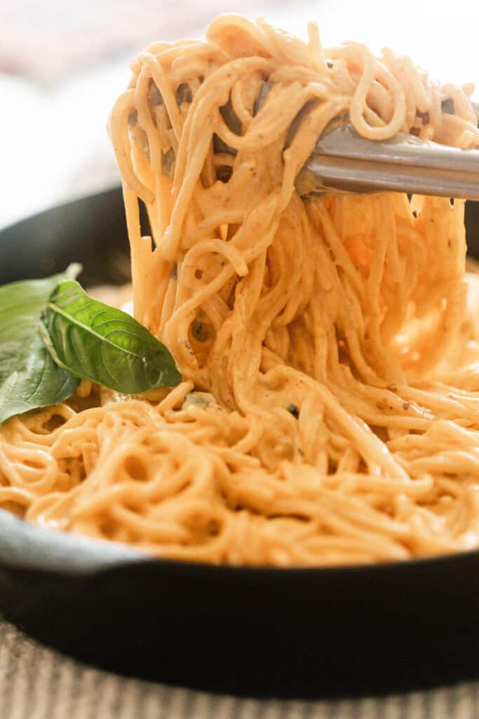 tongs picking up spaghetti noodles  out of a cast iron skillet with basil