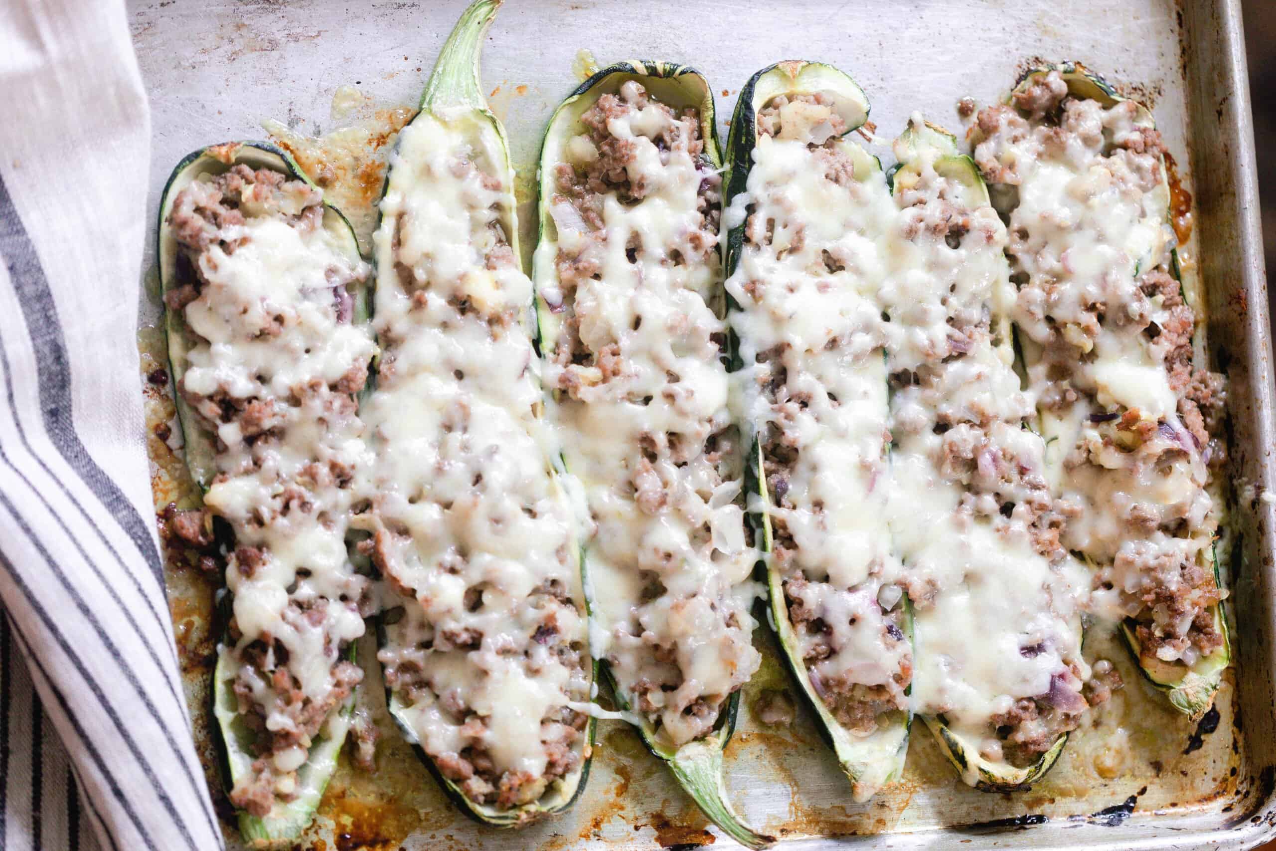 six sausage zucchini boats topped with cheese on a white platter with a gray and white stripped napkin to the left
