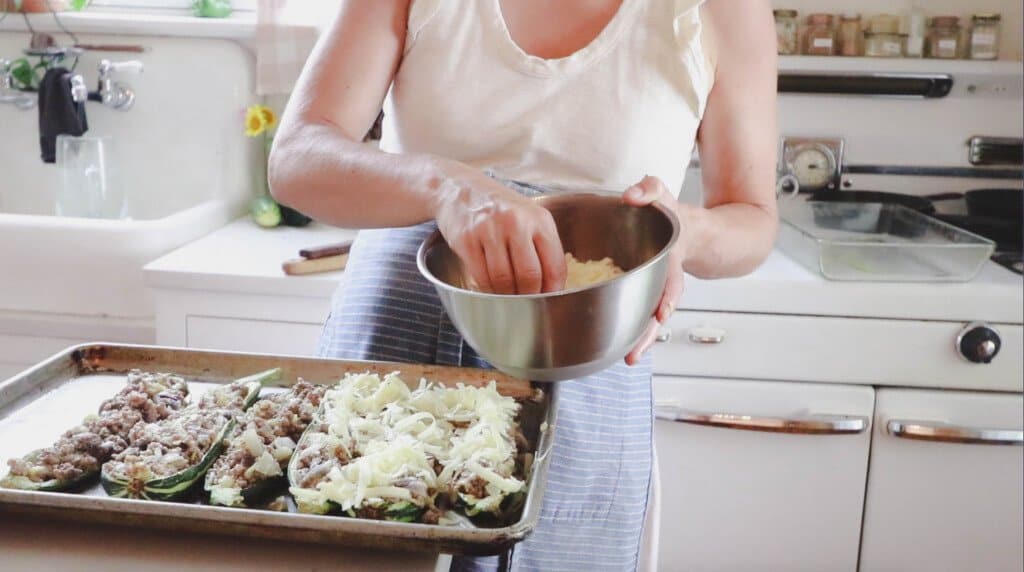 women wearing a white tank top and blue apron sprinting freshly grated cheese to the top of sausage zucchini boats.