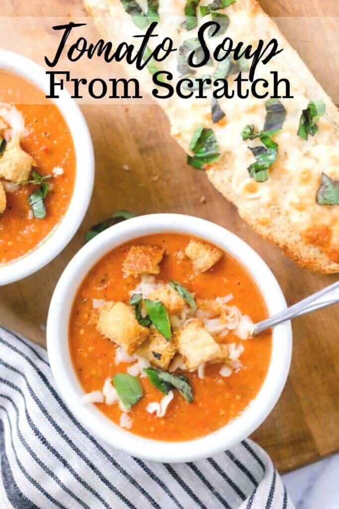 two bowls of tomato soup from scratch with fresh tomatoes topped with fresh basil, croutons, and cheese on a wood cutting board with a loaf of cheesy bread and a white and black stripped towel to the left