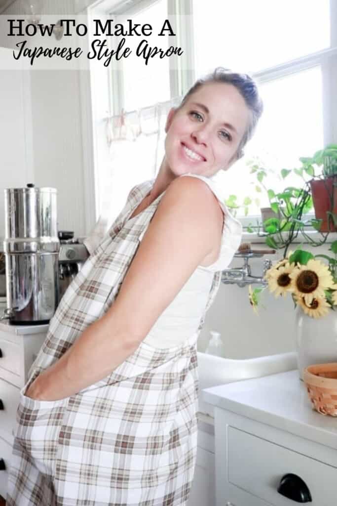 women wearing a plaid Japanese style apron with her hands in the apron pockets. She is staying in a white farmhouse kitchen with a vase of sunflowers in the background