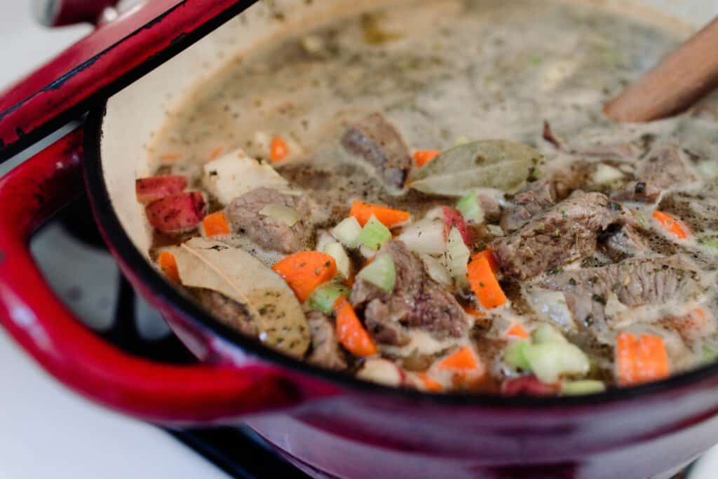red dutch oven with beef stew simmering inside with bone broth, stew meat, carrots, onions, and potatoes
