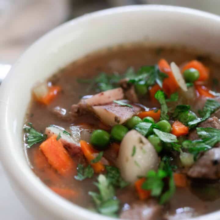 close up picture of beef stew with peas, chunks of carrots, potatoes, and tender beef in a broth and topped with herbs.