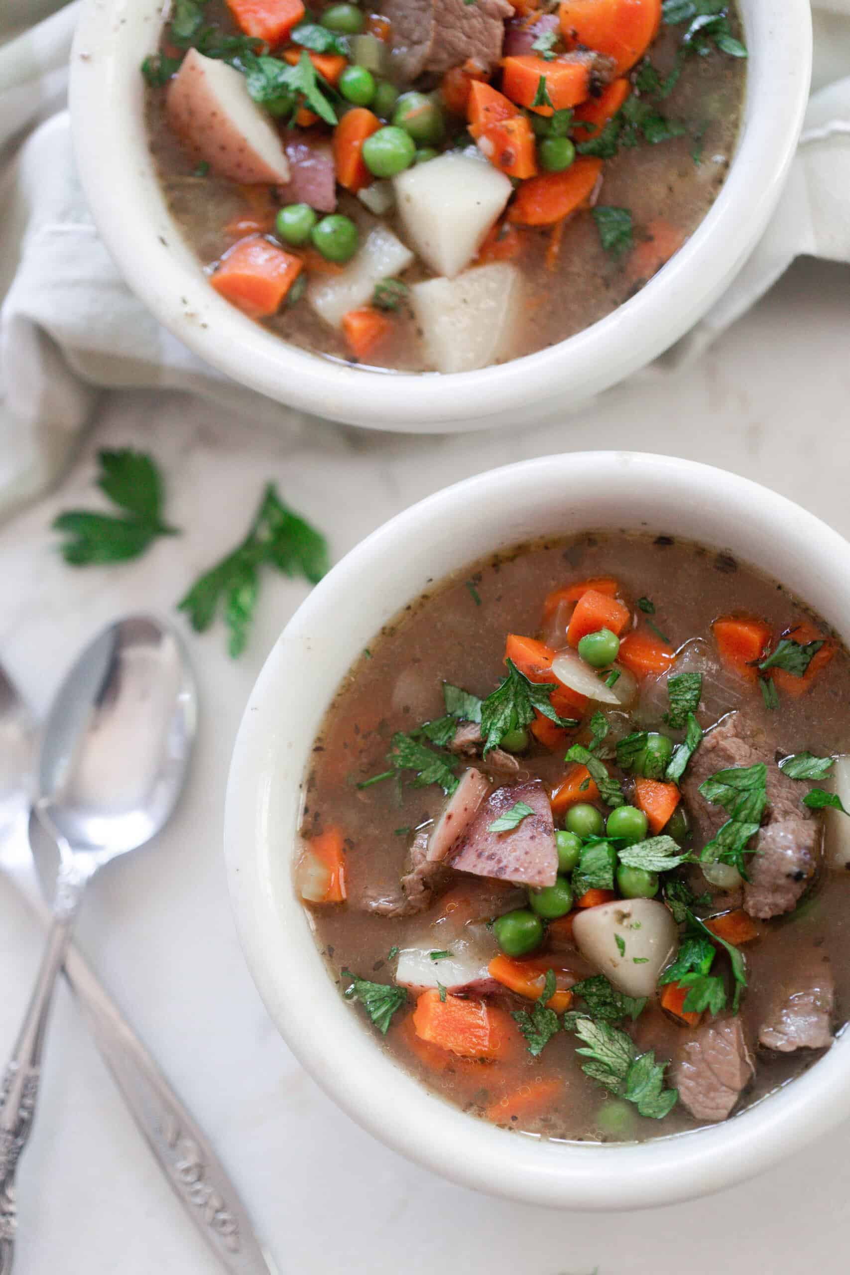 overhead photo of two bowls of beef stew on a white countertop with two spoons to the left of one bowl and a napkin in the background