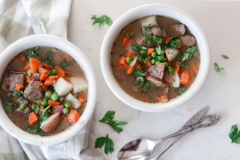 overhead photo of two bowls of stew on a white quartz countertop with a tan and white towel draped over. Two spoons rest in front of the bowls and herbs are sprinkled on the countertop
