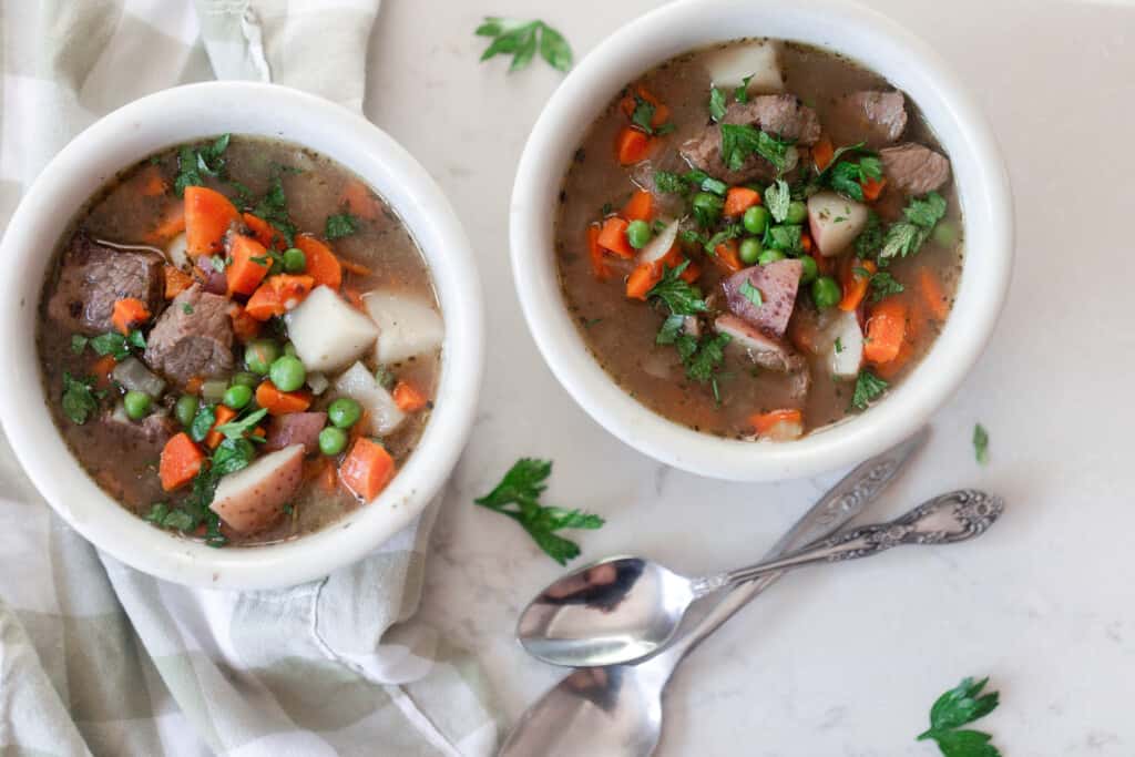 two bowls of beef stew with potatoes, carrots, peas and beef and topped with herbs on a gray countertop