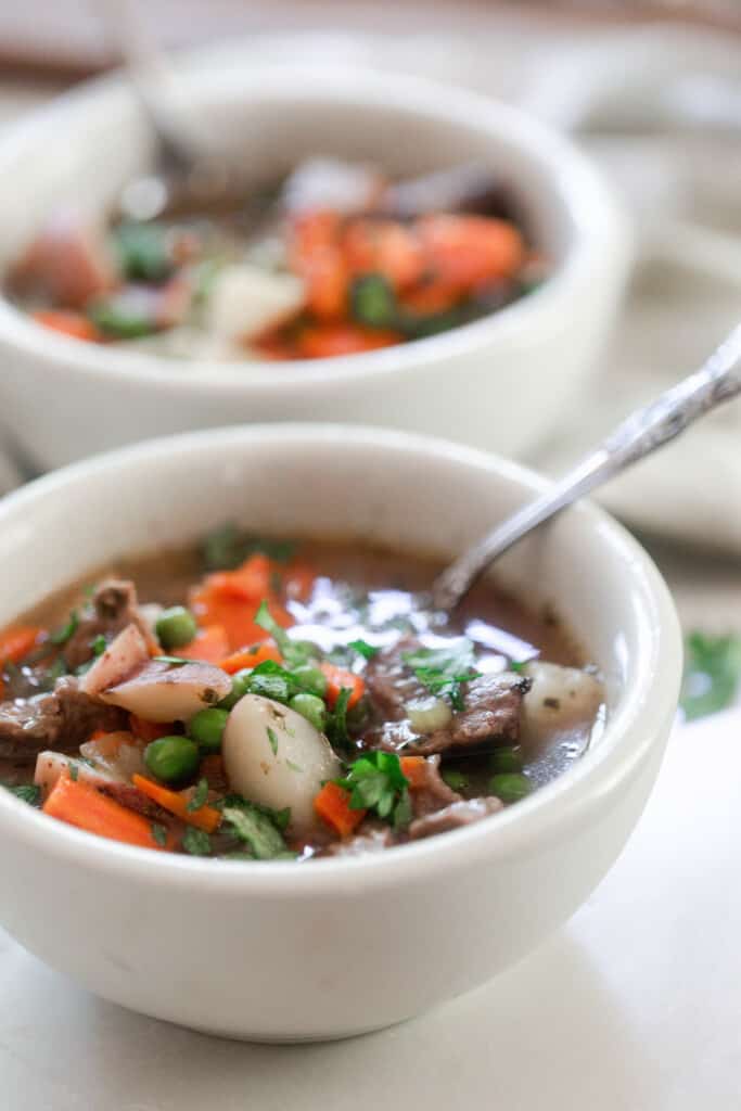 two bowls with one in the front and the other in the background of hearty beef stew packed with potatoes, onions, carrots, and peas