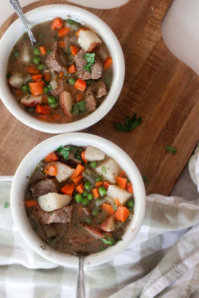 overhead photo of two bowls of beef stew on a wood cutting board with a tan and white checked towel over top
