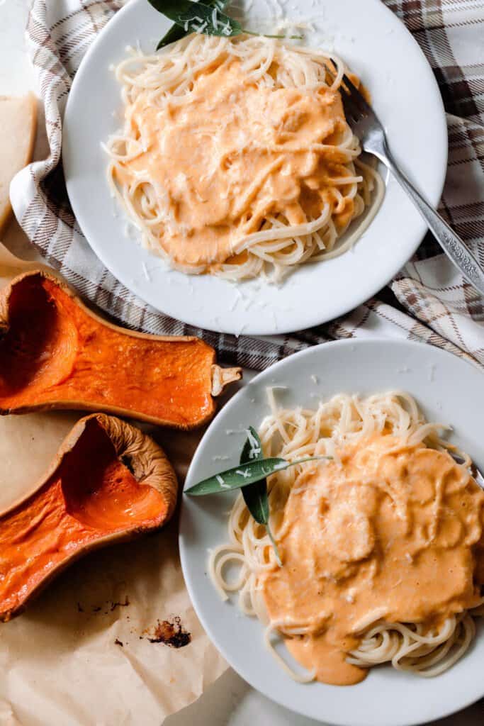 overhead photo of two gray plate with spaghetti topped with butternut squash sauce and garnished with fresh sage. Two halves of baked butternut squash lay on a cutting board to the left