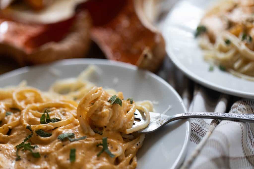 fork with creamy butternut squash pasta wrapped around a fork on a gray plate. Another plate and butternut squash are in the background