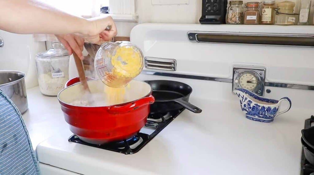 corn in a liquid measuring cup being added to chicken corn chowder in a red dutch oven on a white vintage stove