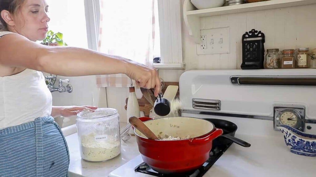 a women adding flour to a red dutch oven on a white antique stove