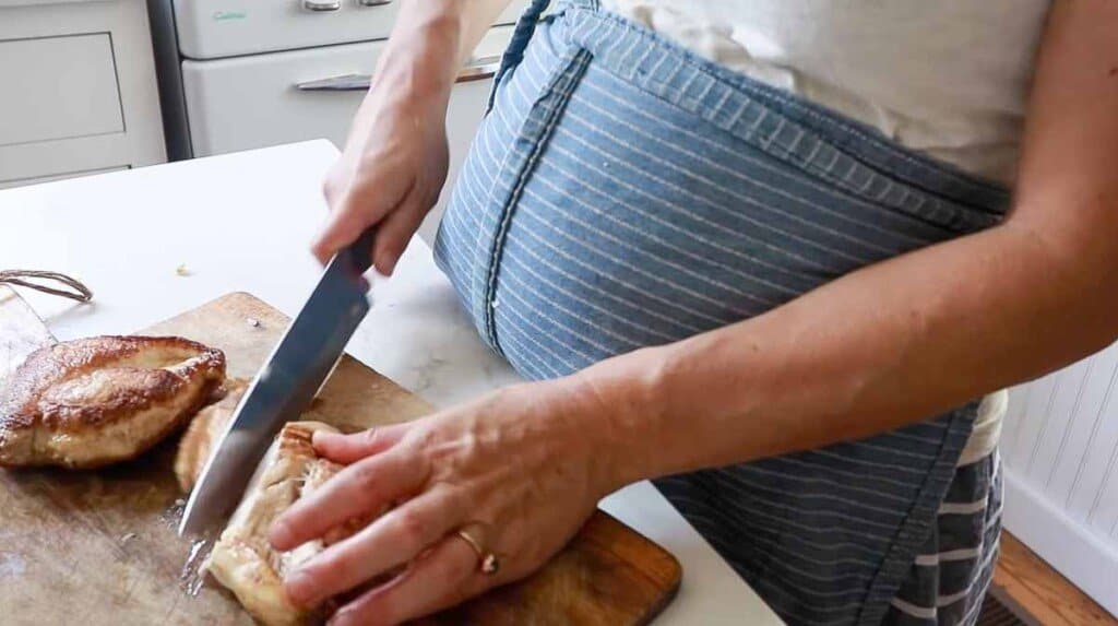 women wearing a blue apron slicing seared chicken on a wood cutting board