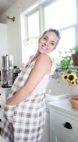 women wearing a plaid Japanese style apron with her hands in the apron pockets. She is staying in a white farmhouse kitchen with a vase of sunflowers in the background